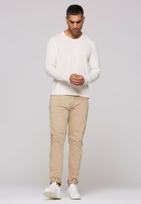 Langärmliges weißes Hemd mit lockerem Schnitt, beigen zulaufenden Hosen und weißen Turnschuhen, stehend vor einem einfarbigen Hintergrund.