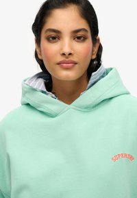 Mintgroene hoodie van zacht fabric, voorzien van een geborduurd "SUPERDRY" logo in koraal. De capuchon is gevoerd met een blauw-wit gestreept patroon.