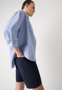 Camisa de botão azul e branca em padrão listrado, com mangas enroladas, combinada com calções escuros de cor azul-marinho. A camisa possui uma bainha curva e um corte descontraído.