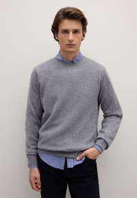 Maglione lavorato a maglia grigio chiaro con colletto rotondo, indossato sopra una camicia a righe blu e bianche con colletto, abbinato a pantaloni scuri.