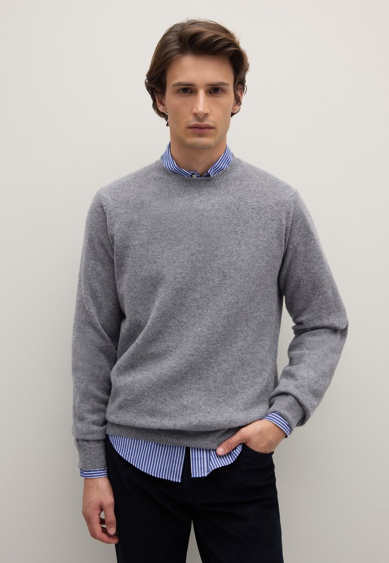Maglione lavorato a maglia grigio chiaro con colletto rotondo, indossato sopra una camicia a righe blu e bianche con colletto, abbinato a pantaloni scuri.