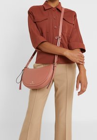 Rosa læder crossbody taske med en buet form, guldfarvet hardware og justerbar rem. Har en glat tekstur og et logo-detallering.