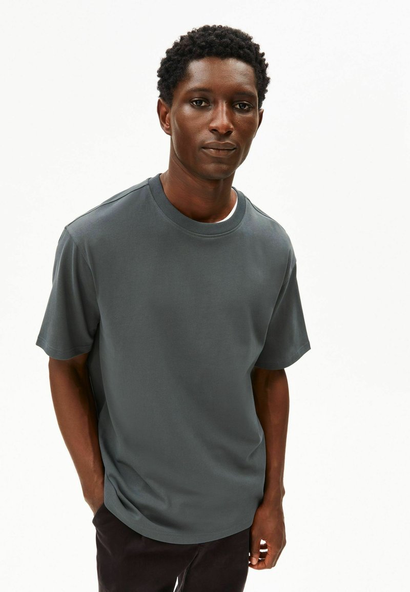 Dunkelgraues Baumwoll-T-Shirt mit Rundhalsausschnitt und kurzen Ärmeln, in entspannter Passform und mit glatter Textur. Das Modell posiert mit den Händen in den Taschen.