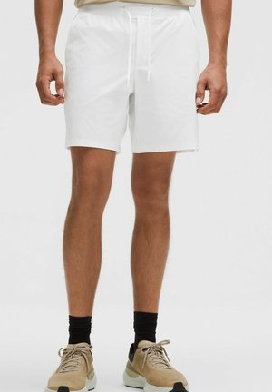 Mann trägt weiße Shorts mit Kordelzug, schwarze Socken und beige Sneakers, steht vor einem schlichten hellen Hintergrund.