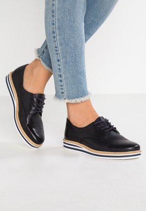 Vrouw in zwarte leren veterschoenen met witte en marineblauwe gestreepte plateauzolen en lichtblauwe spijkerbroek met rafels aan de onderkant.