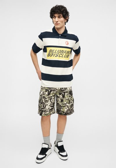 Νεαρός άνδρας στέκεται με τα χέρια πίσω από την πλάτη, φοράει μαύρο-άσπρο ριγέ πόλο με το κείμενο «Billionaire Boys Club» και καμουφλάζ cargo σορτς.