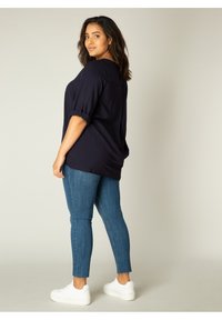 Donkerblauwe oversized blouse met opgerolde mouwen, gecombineerd met strakke blauwe jeans en witte sneakers, met een gladde textuur en casual design.