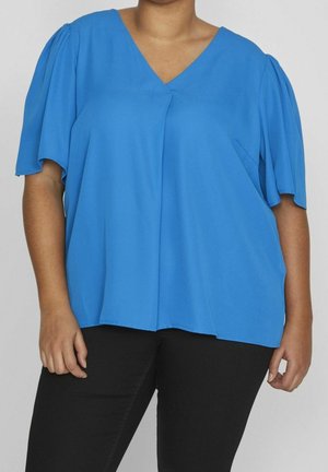 Kobieta ubrana w jasnoniebieską bluzkę z krótkim rękawem i dekoltem w serek oraz czarne spodnie, stojąca na jednolitym szarym tle.