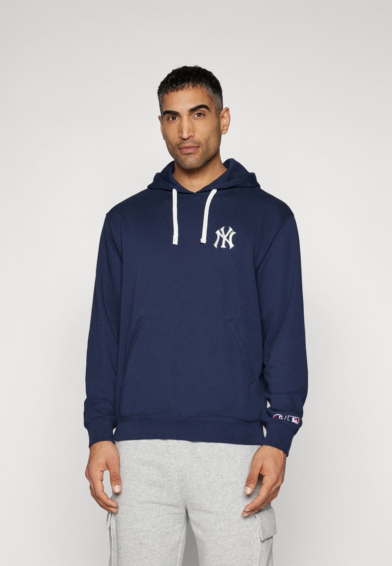 Marinefarbener Kapuzenpullover mit einer vorderen Kängurutasche, weißen Kordeln und einem weißen gestickten Logo auf der linken Brust. Graue Jogginghosen mit Taschen.