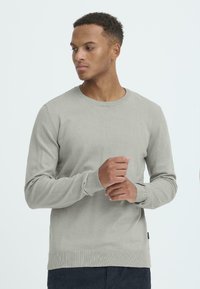 Pulover gri deschis, realizat dintr-un material tricotat moale, prezentând un guler rotund clasic, mâneci lungi și manșete și tiv cu detalii ribbed. Design minimalist.