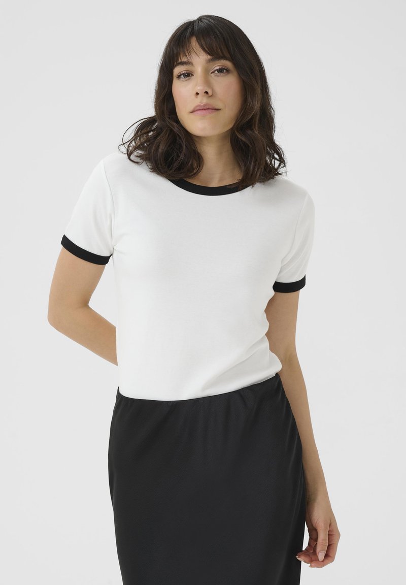 T-shirt blanc à manches courtes avec un col et des poignets côtelés contrastants noirs, fabriqué en tissu lisse. Porté avec une jupe noire.