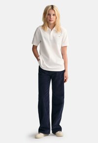 Chemise polo blanche à manches courtes et col, assortie à un pantalon en velours côtelé bleu marine. Le modèle porte des baskets claires, posé contre un fond gris.