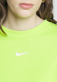Neonově zelená mikina s žebrovaným kulatým výstřihem a bílým logem Nike na hrudi. Látka vypadá měkká a hladká.