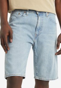 Homme portant un short en jean bleu clair et une chemise beige, montré de la taille jusqu'à mi-cuisse avec les mains détendues sur les côtés.