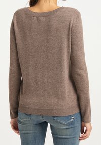 Vrouw in een bruine lange mouwtrui en blauwe jeans, van achteren gezien met haar opgestoken.