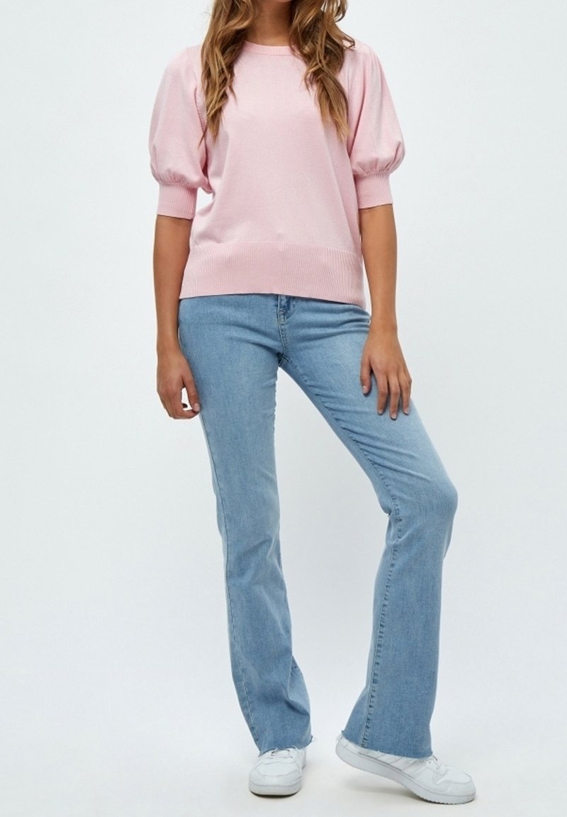 Rosa stickad tröja med puffärmar, kombinerad med ljusblå utsvängda jeans. Modellen har vita sneakers och står mot en ljus bakgrund.