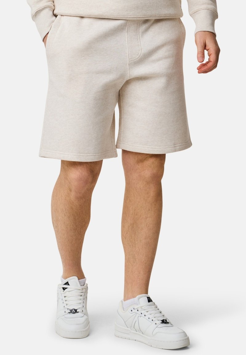 Beige fleece-shorts med elastisk midjeband, sidofickor och en avslappnad passform, parat med vita sneakers med svarta accenter.