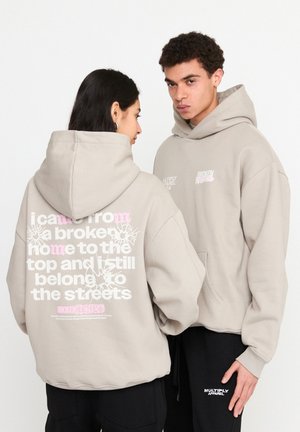 Sweat à capuche en coton beige. Présente un logo sur le devant, un grand texte au dos, et une coupe décontractée. Deux modèles le portent avec des pantalons contrastants.