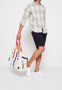 Witte canvas tas met rode, blauwe en gele handvaten; heeft twee zijzakken en een ritsvak in het midden. Model gekleed in een geruit shirt en shorts.