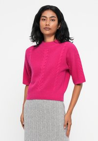 Pull en tricot rose à manches courtes avec des motifs en torsades, présentant un col rond côtelé et un ourlet cropped. Porté avec une jupe grise côtelée.