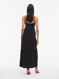 Rochie maxi neagră cu bretele subțiri, design în straturi și textură moale. Asortată cu sandale cu toc roșii, evidențiind o siluetă simplă și elegantă.