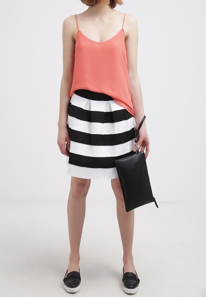 Femme portant un caraco corail, une jupe rayée noire et blanche, des mocassins noirs, tenant une pochette noire pour poignet, debout devant un fond uni.