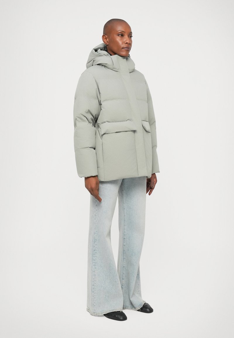 Veste rembourrée gris clair avec une capuche, fermeture éclair centrée et deux poches avant. Associée à un jean bleu délavé à jambes larges et des chaussures noires.