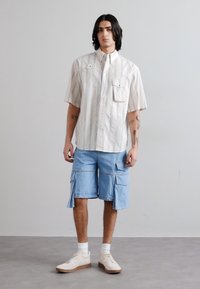 Camicia a maniche corte con colletto a righe verticali blu e bianche, abbinata a pantaloni cargo blu chiaro con tasche frontali e zip.