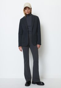 Woman wearing a beige cap, dark gray turtleneck, blazer, matching pants, and black chunky shoes, standing against a plain white wall.