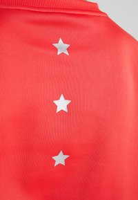 Maglietta sportiva rossa con tre stelle argentate in rilievo allineate verticalmente sul retro. Realizzata in tessuto texturizzato per una migliore traspirabilità.