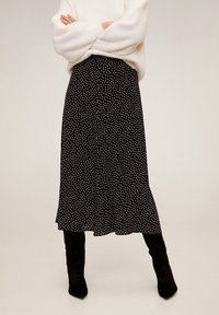 Vrouw in een zwarte midi-jurk met witte abstracte stippen, gecombineerd met een witte fuzzy trui en zwarte kniehoge laarzen.