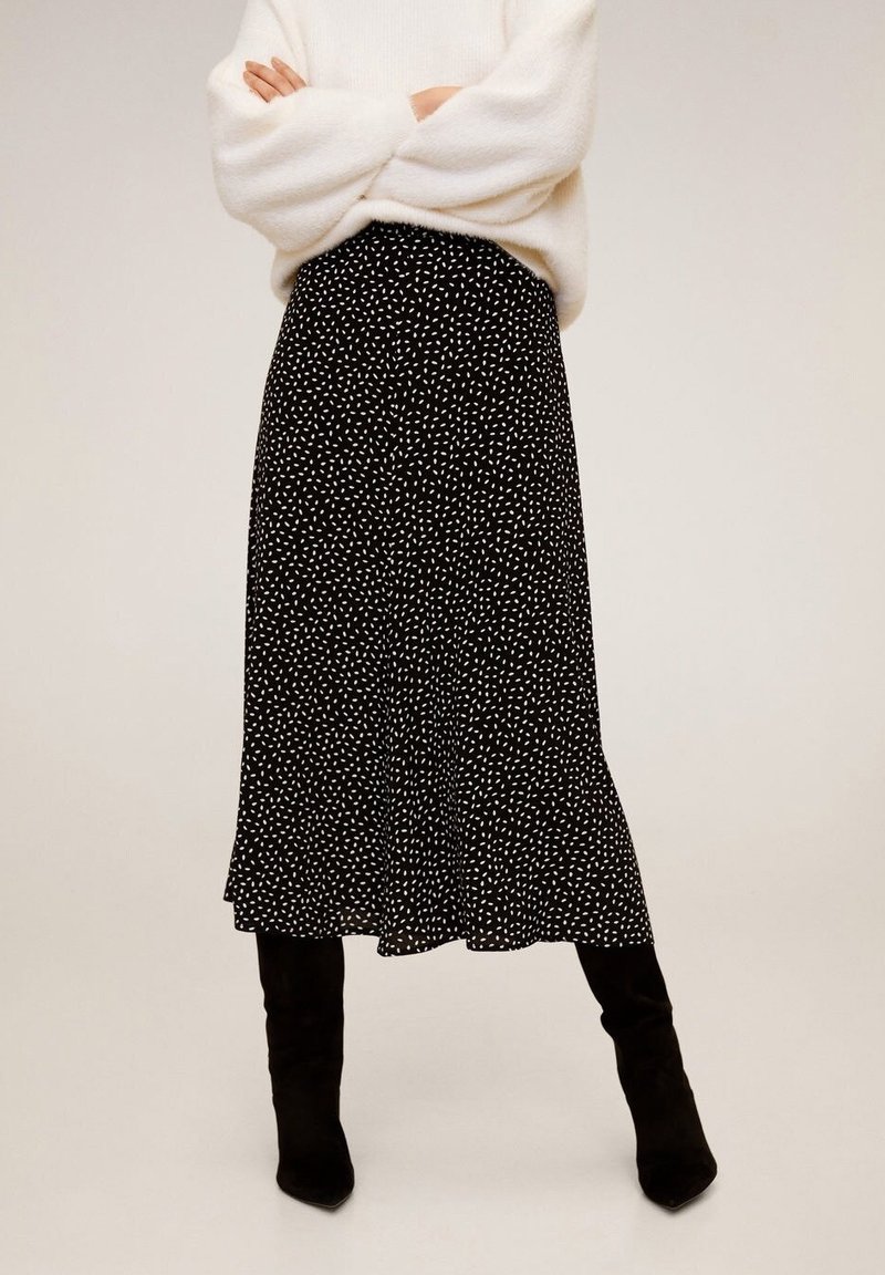 Vrouw in een zwarte midi-jurk met witte abstracte stippen, gecombineerd met een witte fuzzy trui en zwarte kniehoge laarzen.