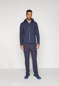 Tamsiai mėlyna lengva striukė su gaubtu, turinti priekinį užtrauktuką ir prigludusio dizaino, derinama su tamsiai mėlynomis kelnėmis ir mėlynais sportiniais bateliais.