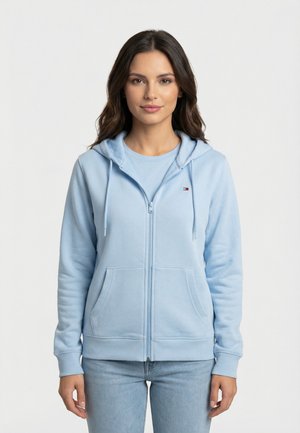 Donna con lunghi capelli castani che indossa una felpa con cappuccio azzurra con cerniera e jeans, in piedi davanti a uno sfondo bianco uniforme.