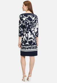 Navyblaues Blumen-Kleid mit weißen Konturen, drei Viertel-Ärmeln und einem figurbetonten Design mit einem gemusterten Saum. Kombiniert mit weißen Blockabsätzen.