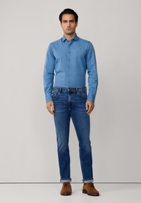 Lichtblauw overhemd met knopen, donkerblauwe jeans, bruine veterschoenen. Gladde stof, getailleerde pasvorm, opgerolde mouwen en minimalistische merkmale.