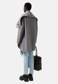 Šedý vlněný šál přehozený přes prošívanou šedou bundu; světle modré džíny a robustní černé boty; drží černou tašku.