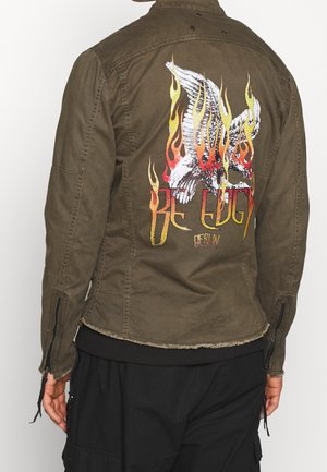 Oliwkowa kurtka z grafiką płonącego białego ptaka i napisem „Be Edgy Berlin” na plecach, noszona z czarnymi spodniami.