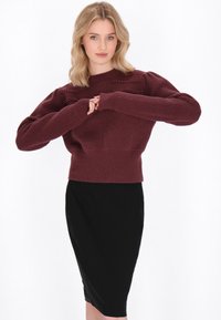 Burgundijas adīts džemperis ar ribotām malām un teksturētām akcentiem plecos, valkāts virs pieguļoša melna svārku. Mīksta, nedaudz pūkaina tekstūra.