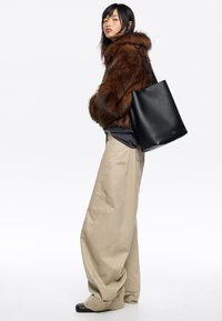 Brązowy futrzany płaszcz, czarna skórzana torba na ramię, szary top, beżowe, oversize'owe spodnie, czarne buty. Model stoi na neutralnym tle.