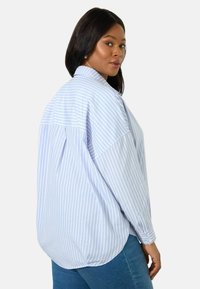 Camicetta button-up a righe light blue con una vestibilità rilassata, maniche lunghe e orlo curvo. Il tessuto appare morbido con una consistenza croccante.