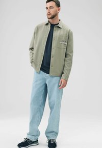 Light green button-up jacket with chest pocket, worn over a navy t-shirt, paired with loose light blue jeans and black sneakers.