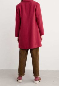 Abrigo rojo texturizado con un cuello ancho y corte recto. Combinado con pantalones de pana verde oliva y zapatos marrón claro.