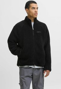 Chaqueta de forro polar negra con cierre de cremallera, cuello alzado y mangas raglán. Cuenta con un interior suave y un sutil logo en el pecho.