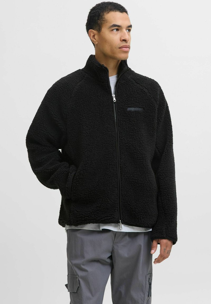 Chaqueta de forro polar negra con cierre de cremallera, cuello alzado y mangas raglán. Cuenta con un interior suave y un sutil logo en el pecho.