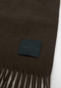 Rjava volna šal z resicami, ki ima temno zelen kvadratni oznako z napisom "KLASIČNO. MODERNO. VAŠE." Tekstura mehka in prijetna na otip.