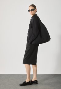 Svart överdimensionerad väska, ribbad svart blus, knälång svart kjol och glänsande svarta loafers. Modellen bär svarta solglasögon.