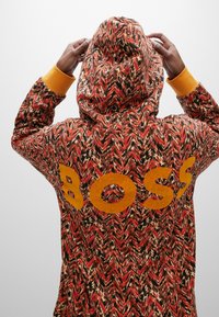 Sudadera con capucha con un patrón en zigzag rojo, naranja y negro. Presenta un prominente logo amarillo "BOSS" en la parte posterior y puños amarillos.