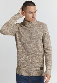 Lys brun strikket genser med høy ribbestrikket krage, teksturert mønster og en merketlapp nederst til venstre. Brukt med blå jeans.