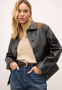 Musta nahkatakki, jossa on kaulus, viisi etunappia ja näkyvät ompeleet. Päällä beige villapaita ja siniset farkut, joissa on ruskea vyö.